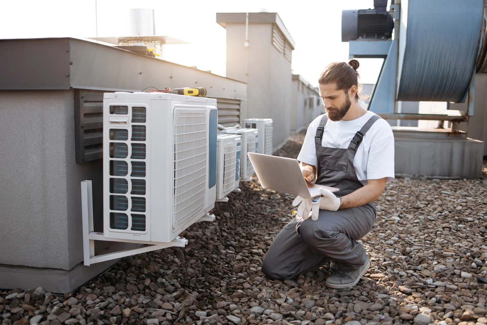 Technicien en climatisation travaillant sur un toit, agenouillé près d'unités de climatisation extérieures, utilisant un ordinateur portable pour diagnostic ou maintenance