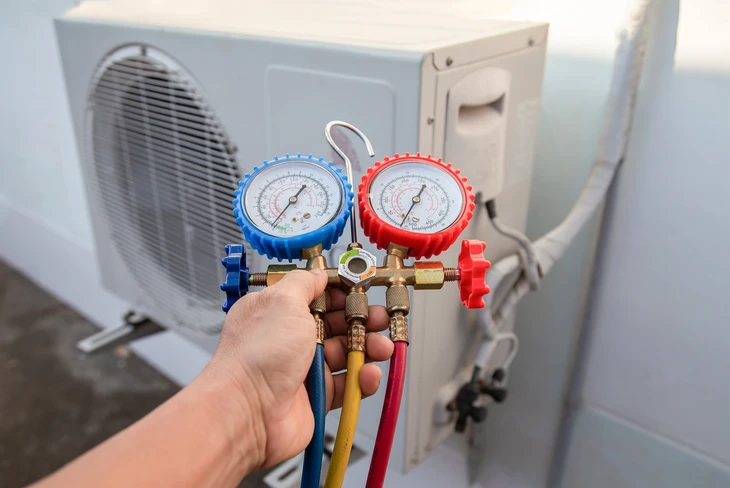 Technicien en tenue de travail professionnelle effectuant un contrôle de maintenance sur une unité de climatisation intérieure (split mural) ou un système de réfrigération professionnel. Le technicien utilise des outils et un multimètre pour vérifier les fluides frigorigènes et la performance du système. L'environnement est celui d'un bâtiment commercial ou professionnel. La scène illustre concrètement une intervention de maintenance préventive sur un équipement de climatisation.